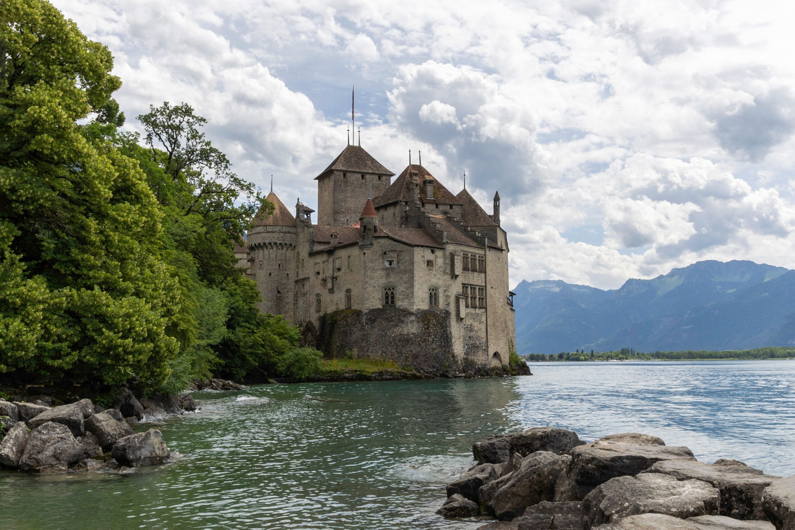 Scenic view of Château de Chillon by the shores of Lake Geneva, surrounded by mountains and lush greenery.